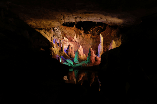 A brightly lit underground cave with colorful stalagmites, stalactites, and their reflections in still water.