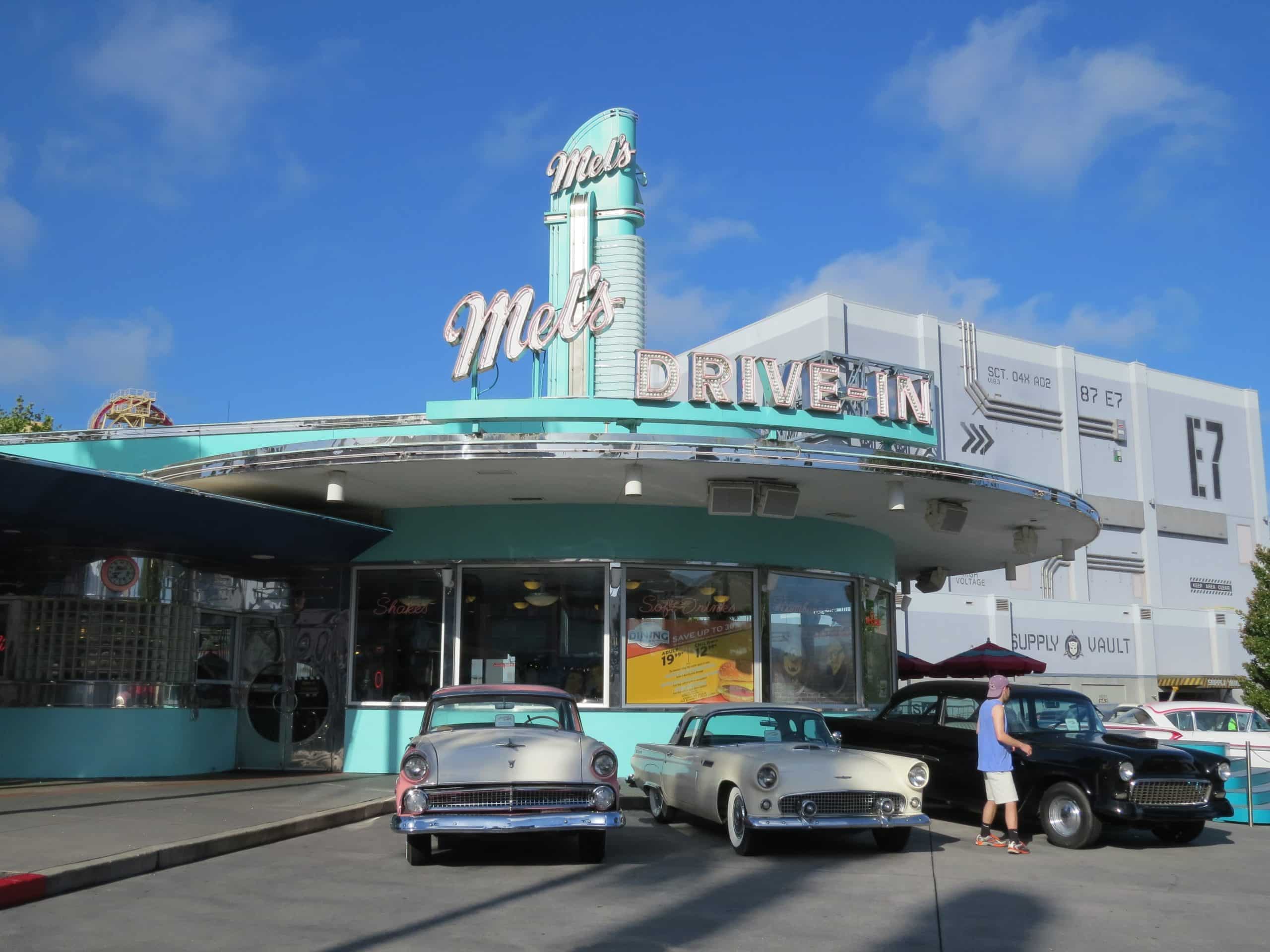 Retro-style Mel's Drive-In diner with classic cars parked in front under a bright blue sky.
